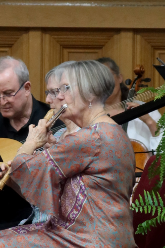 A woman playing flute, with a woman playing recorder and a man playing theorbo behind her.